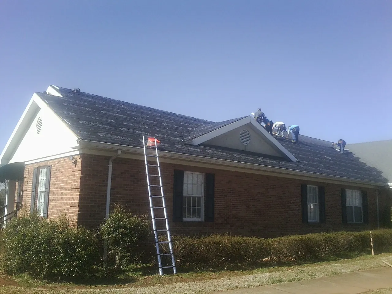 Crew tearing shingles off a brick ranch roof with a ladder set — Metal Roof Repair work by our crew in Fischer